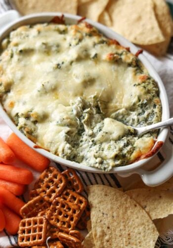 A spoon scooping spinach and artichoke dip surrounded by carrots, pretzels, and tortilla chips
