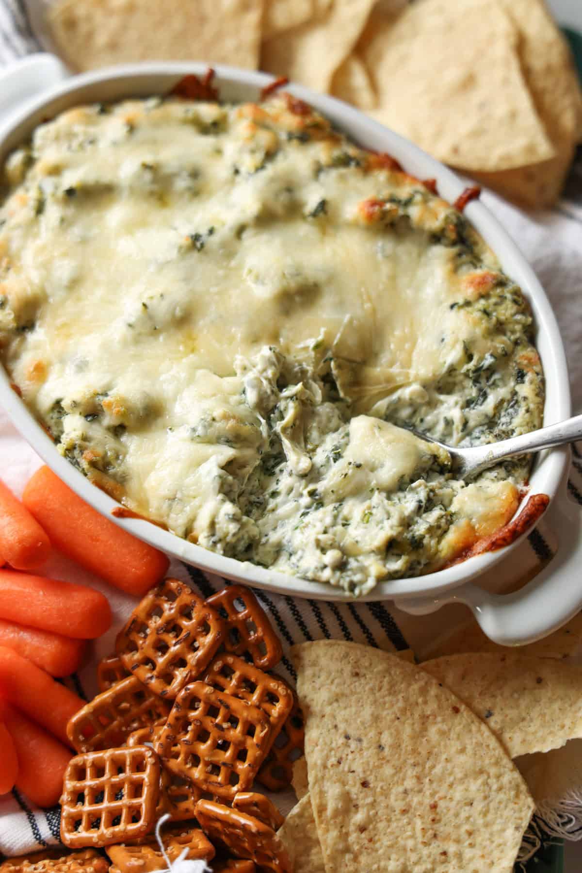 A spoon scooping spinach and artichoke dip surrounded by carrots, pretzels, and tortilla chips