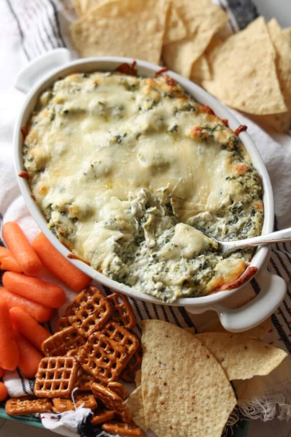 A platter of spinach and artichoke dip baked in a white ceramic dish with a spoon