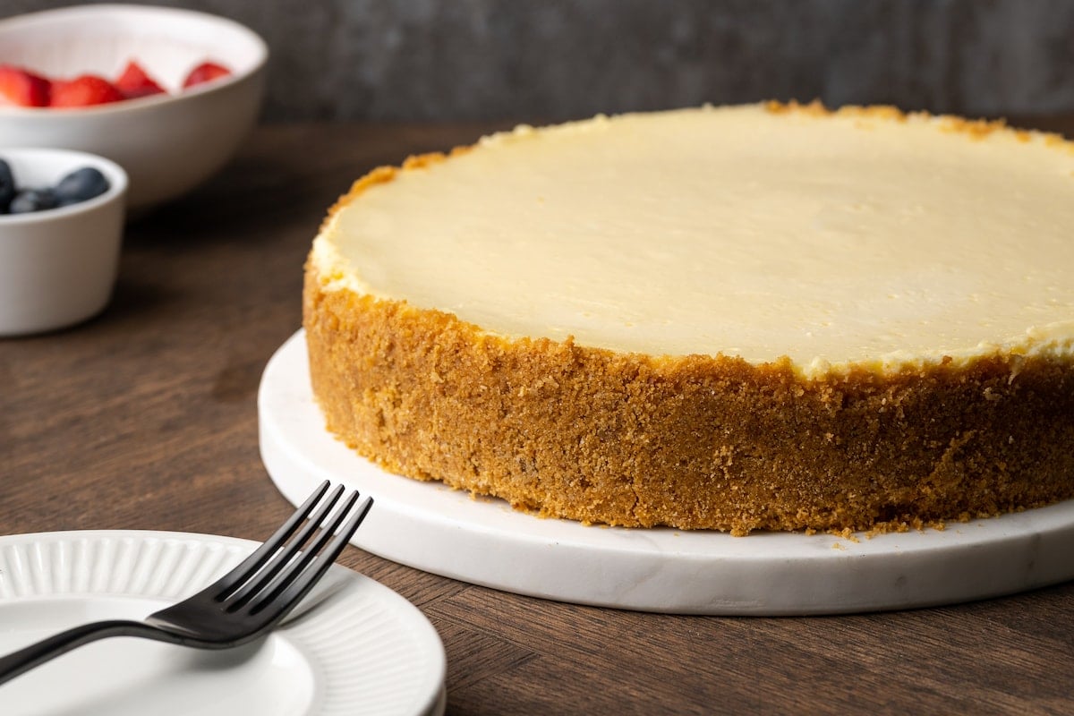 A whole sour cream cheesecake on a white plate, next to a plate with a fork.
