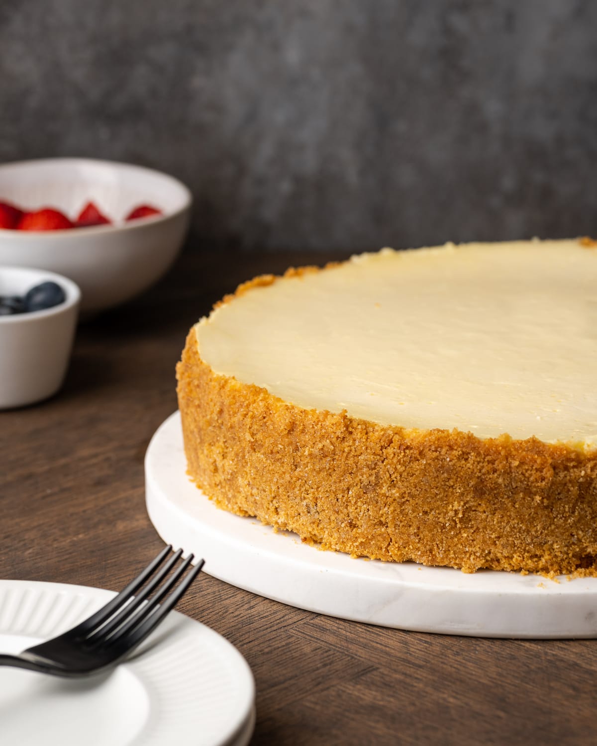 A whole sour cream cheesecake on a white plate, next to a plate with a fork.