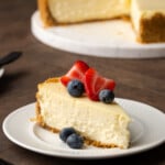 A slice of sour cream cheesecake garnished with fresh berries on a white plate, with the full cheesecake in the background.