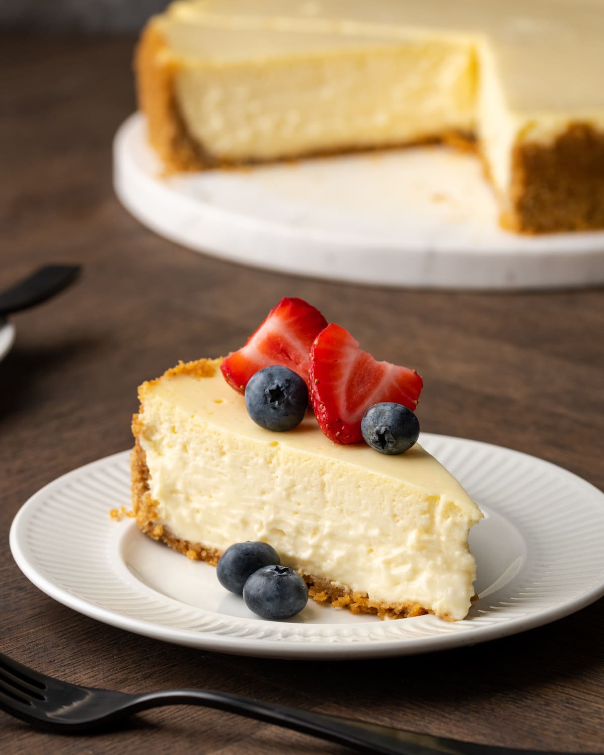 A slice of sour cream cheesecake garnished with fresh berries on a white plate, with the full cheesecake in the background.