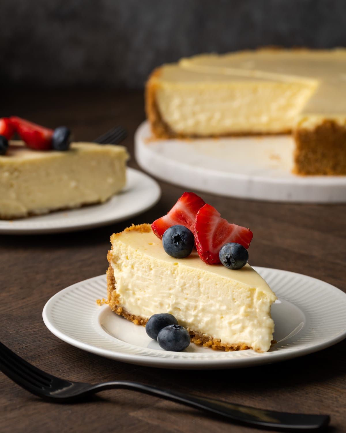 A slice of sour cream cheesecake garnished with fresh berries on a white plate, with the full cheesecake in the background.