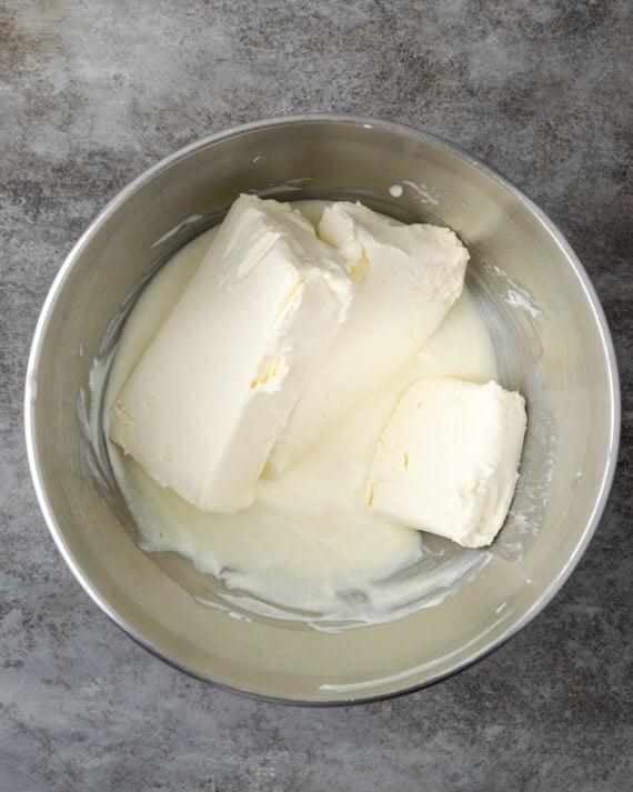 Cream cheese blocks are added into a bowl with sour cream and sugar for cheesecake batter.