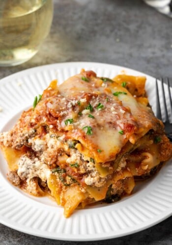 A slice of Instant Pot lasagna on a plate with a fork, next to a glass of white wine.