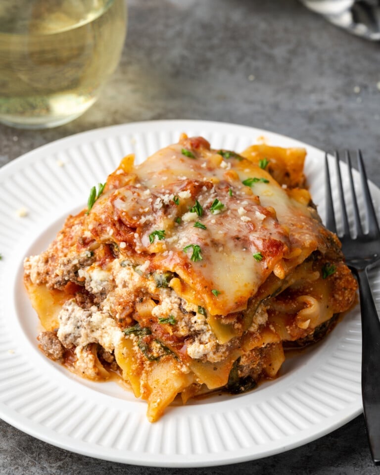 A slice of Instant Pot lasagna on a plate with a fork, next to a glass of white wine.