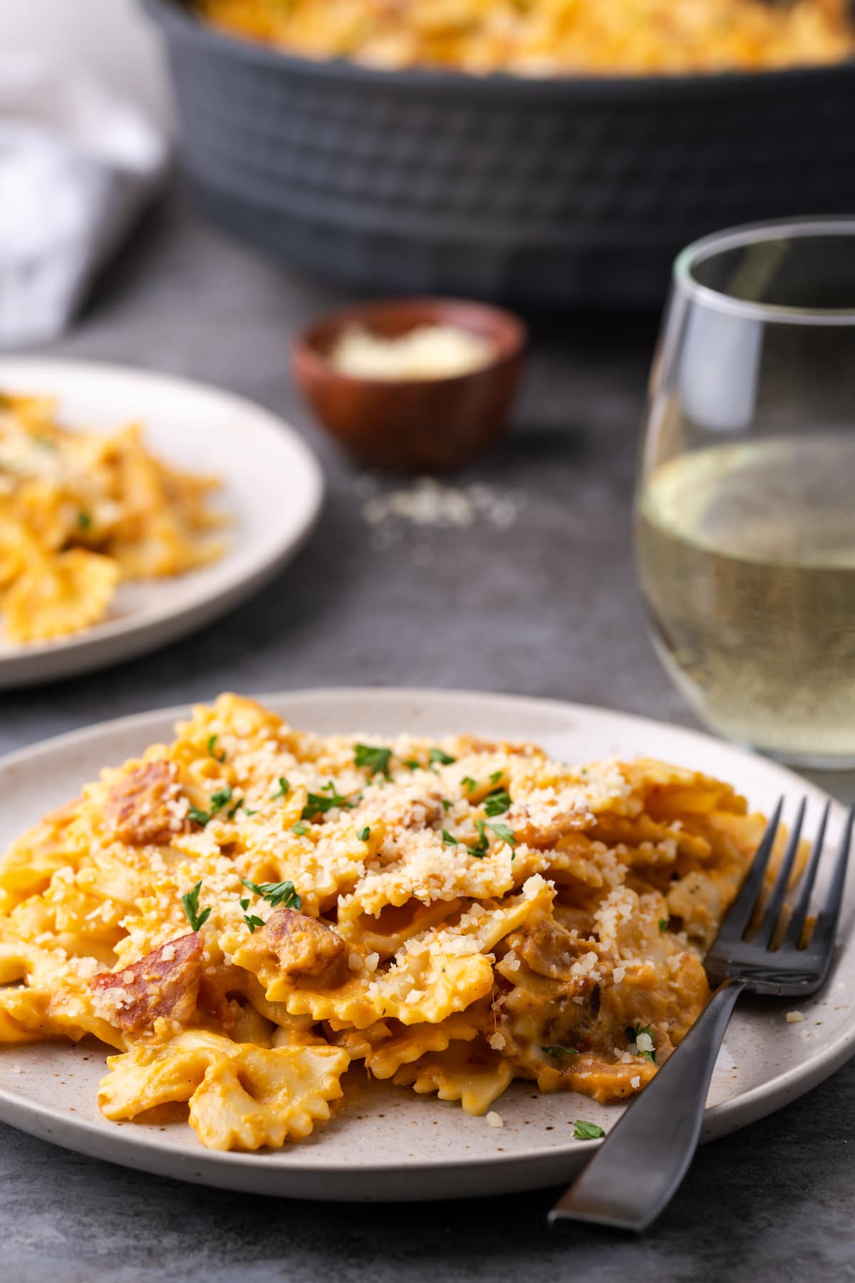 A serving of pumpkin pasta with bacon on a plate next to a fork, with a glass of white wine in the background.