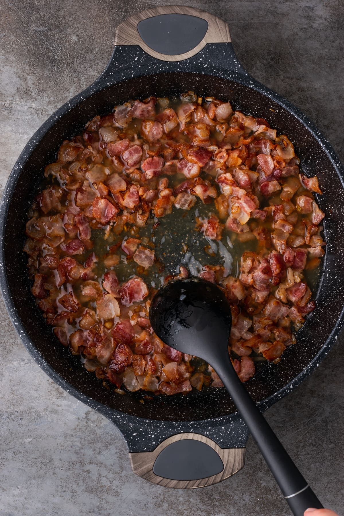 A plastic spoon scrapes the bottom of a pan filled with caramelized onions and bacon, deglazed with broth and white wine.