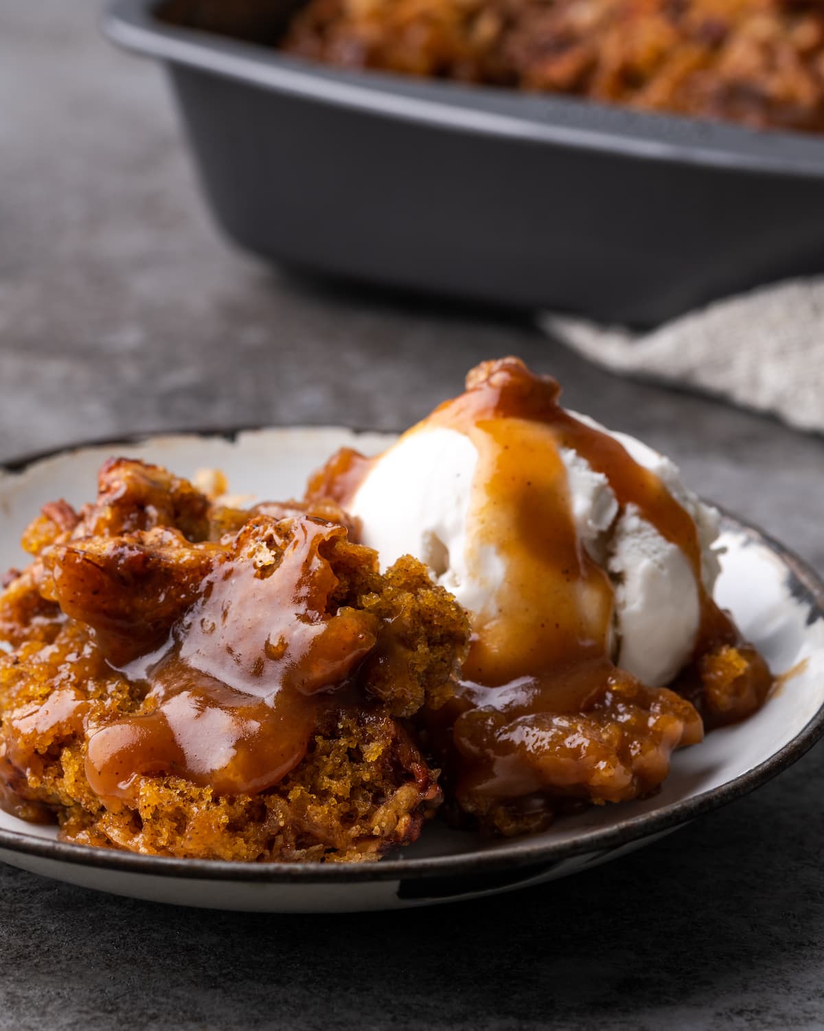 A serving of pumpkin cobbler on a white plate topped with a scoop of vanilla ice cream and caramel sauce.