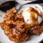 A serving of pumpkin cobbler on a white plate topped with a scoop of vanilla ice cream and caramel sauce.
