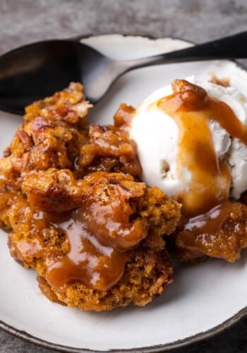 A serving of pumpkin cobbler on a white plate topped with a scoop of vanilla ice cream and caramel sauce.