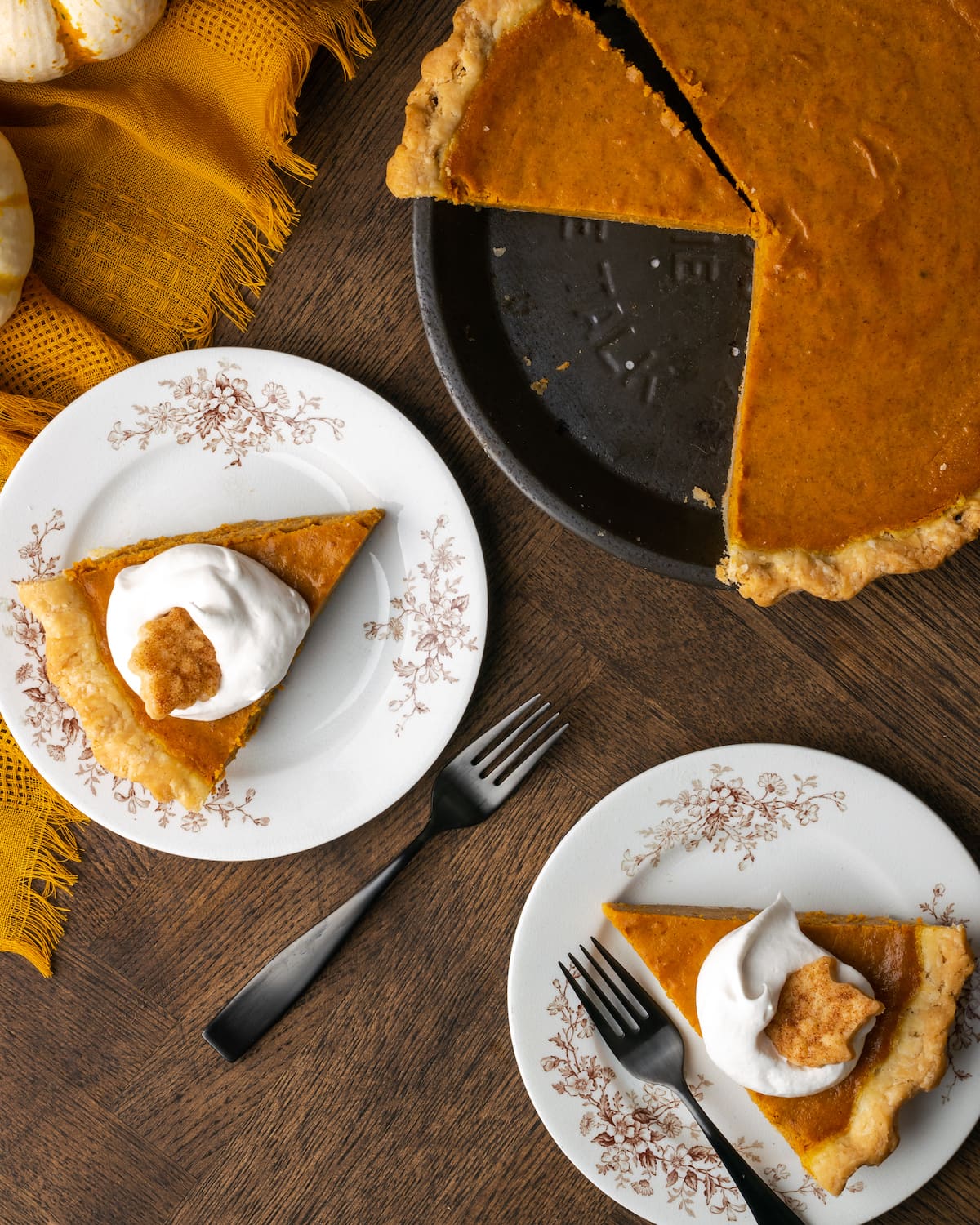 Overhead view of two slices of pumpkin pie on plates, topped with whipped cream, next to a pumpkin pie with slices missing.