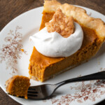 A slice of pumpkin pie on a plate topped with whipped cream and a sugar cookie, with a forkful of pie missing.