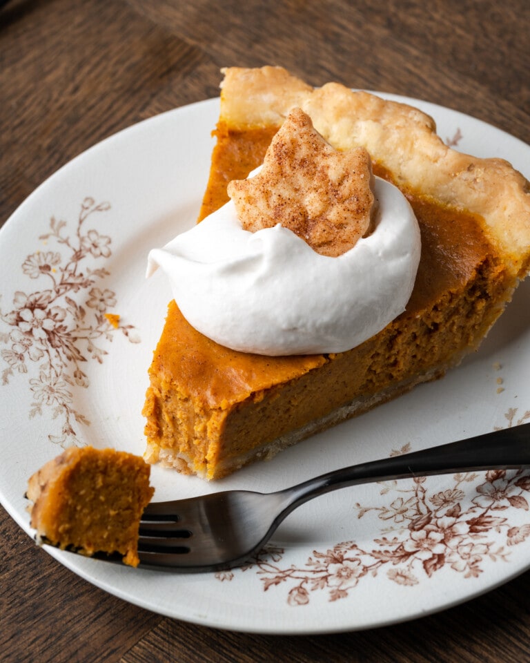 A slice of pumpkin pie on a plate topped with whipped cream and a sugar cookie, with a forkful of pie missing.