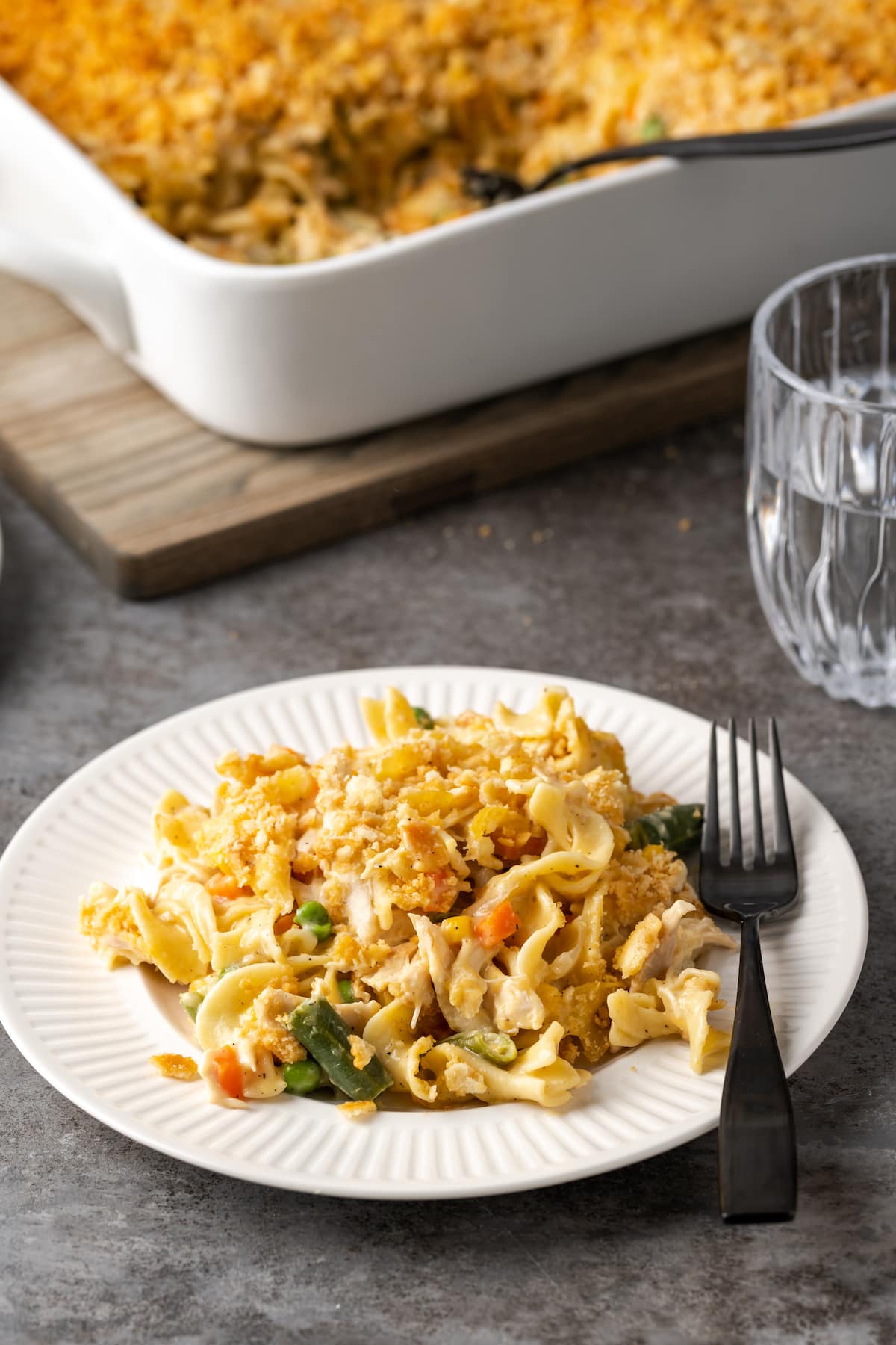 A serving of chicken noodle casserole on a white plate with a fork, with the casserole in the background.