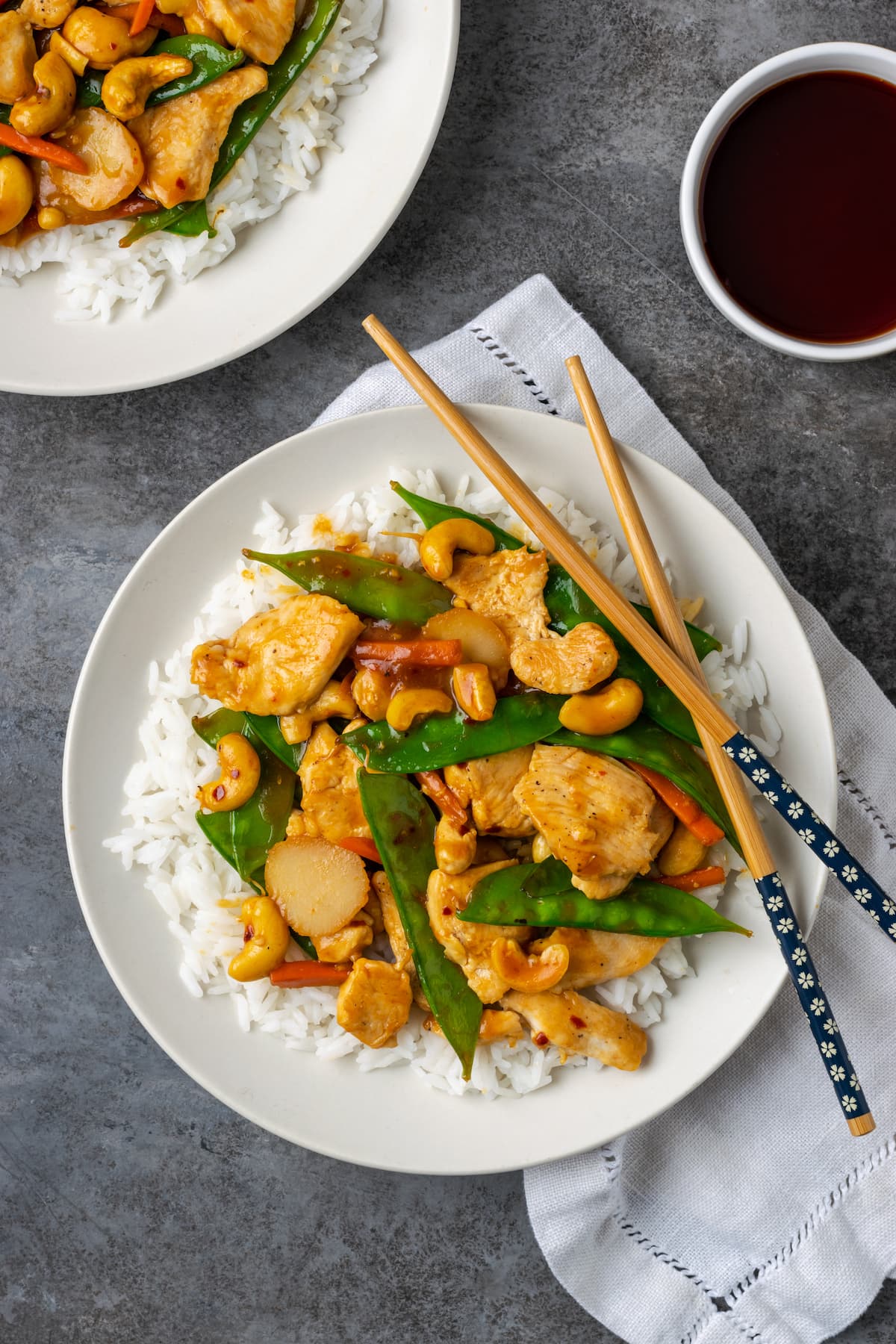 Overhead view of cashew chicken served over a bed of jasmine rice on a plate, next to chop sticks and a bowl of soy sauce.