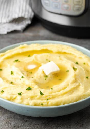 Creamy Instant Pot mashed potatoes in a bowl topped with two pats of butter and chives, with the Instant Pot in the background.