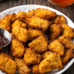 Crispy air fryer popcorn chicken on a white place next to a ramekin of BBQ sauce for dipping.