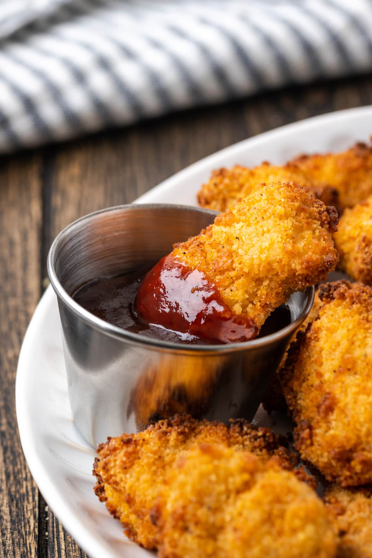 A piece of popcorn chicken partially dipped into a ramekin of BBQ sauce, on a plate with more popcorn chicken pieces.