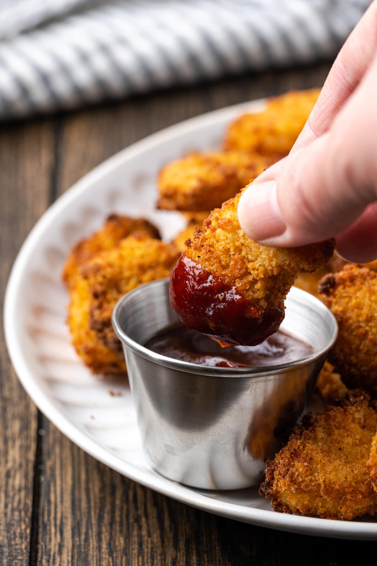 A hand holding a piece of popcorn chicken partially dipped into a ramekin of BBQ sauce, on a plate with more popcorn chicken pieces.