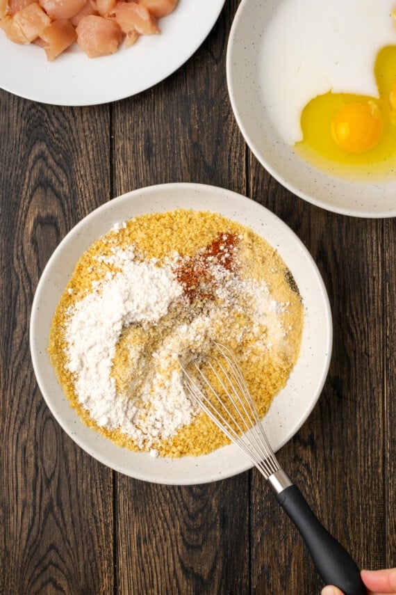 The ingredients for the panko breadcrumb coating are whisked together in a white bowl, next to a bowl of raw chicken pieces and a bowl with eggs.