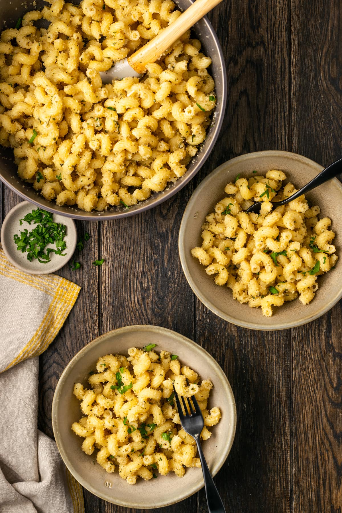 Overhead view of two bowls of garlic bread pasta next to a large serving bowl of pasta with a wooden spoon.