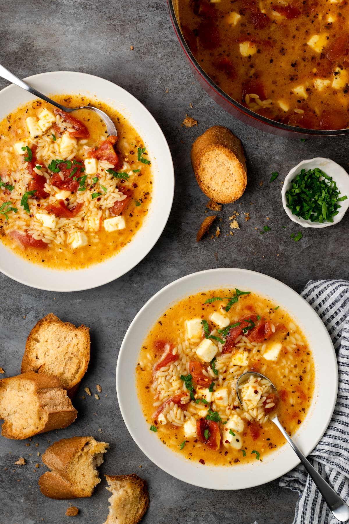 Overhead view of two bowls of Greek tomato feta soup with spoons, next to slices of bread.