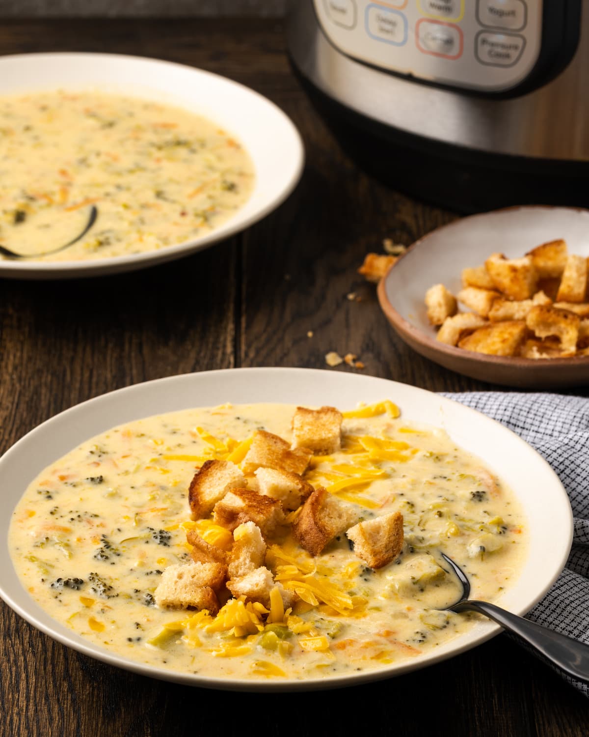 A bowl of instant pot broccoli cheddar soup topped with croutons with a spoon, with a second bowl of soup next to a bowl of croutons in the background.