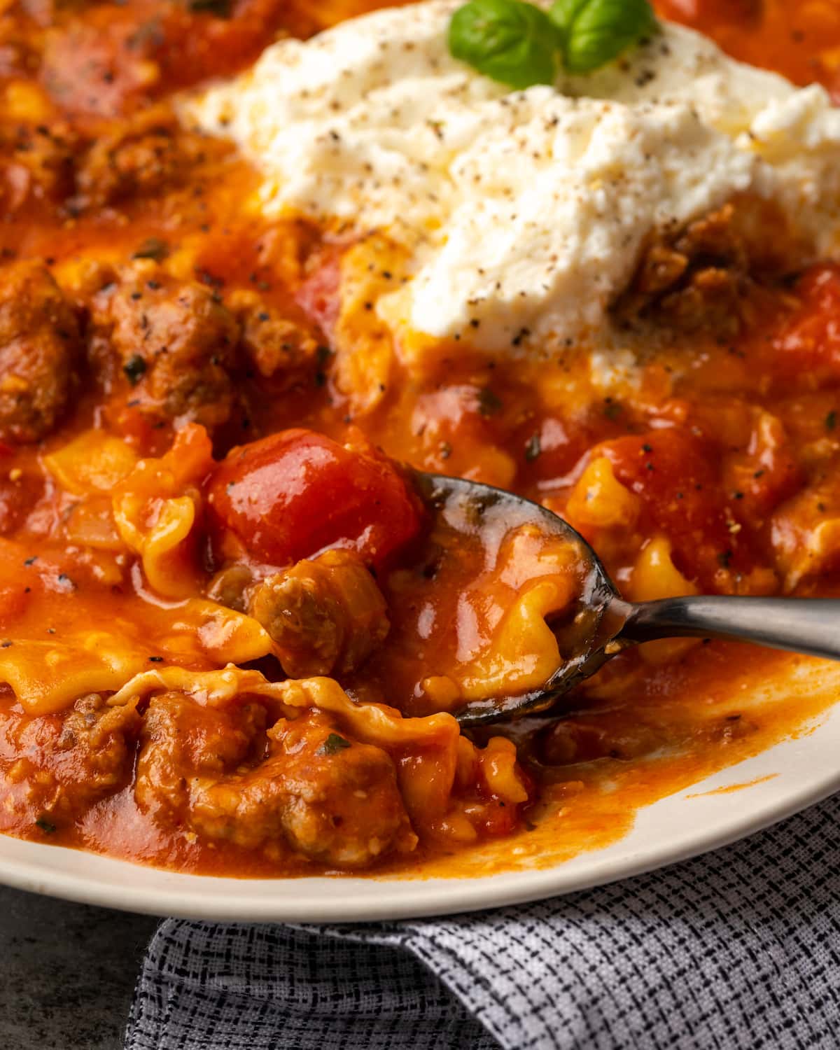 A bowl of lasagna soup topped with ricotta cheese and basil leaves, with a spoon.