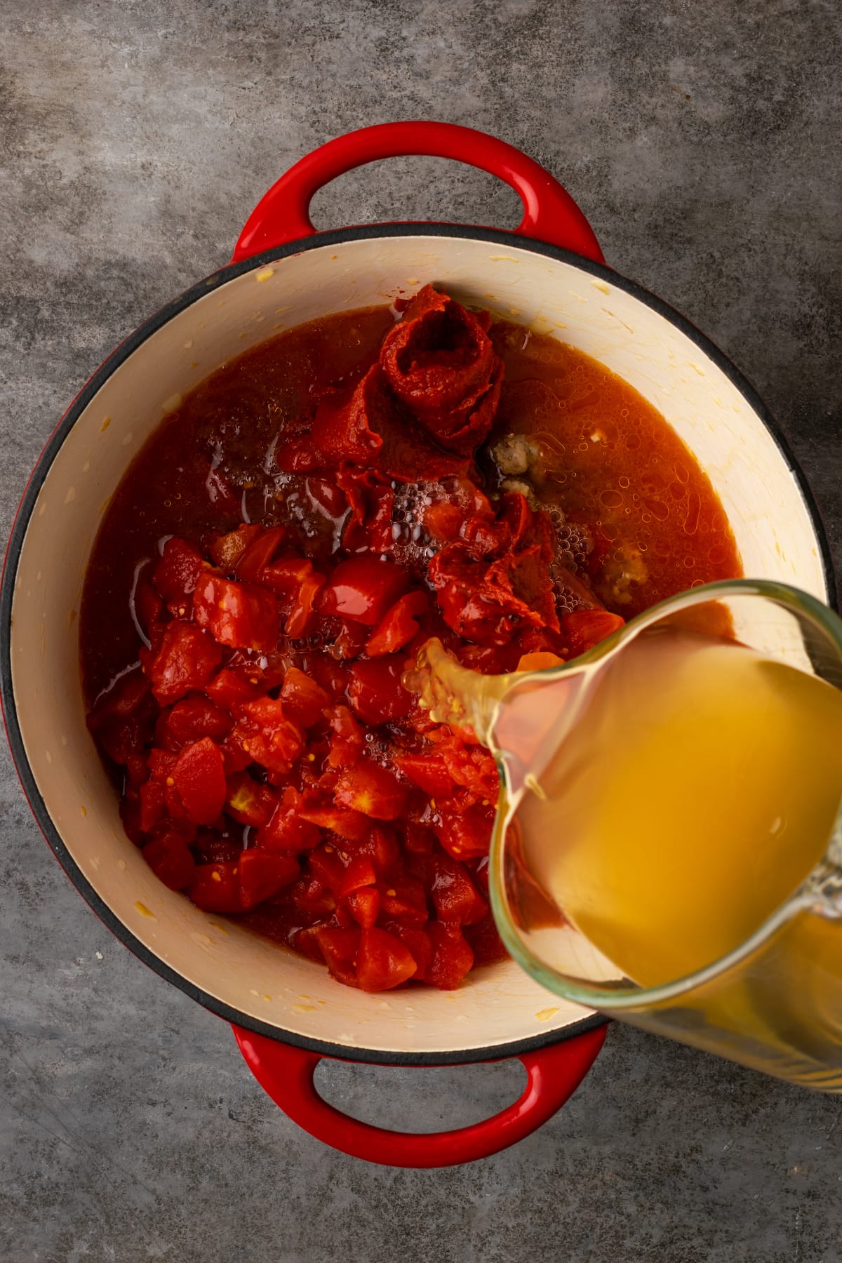 Chicken broth is poured into a large pot with diced tomatoes, tomato paste, and ground sausage.