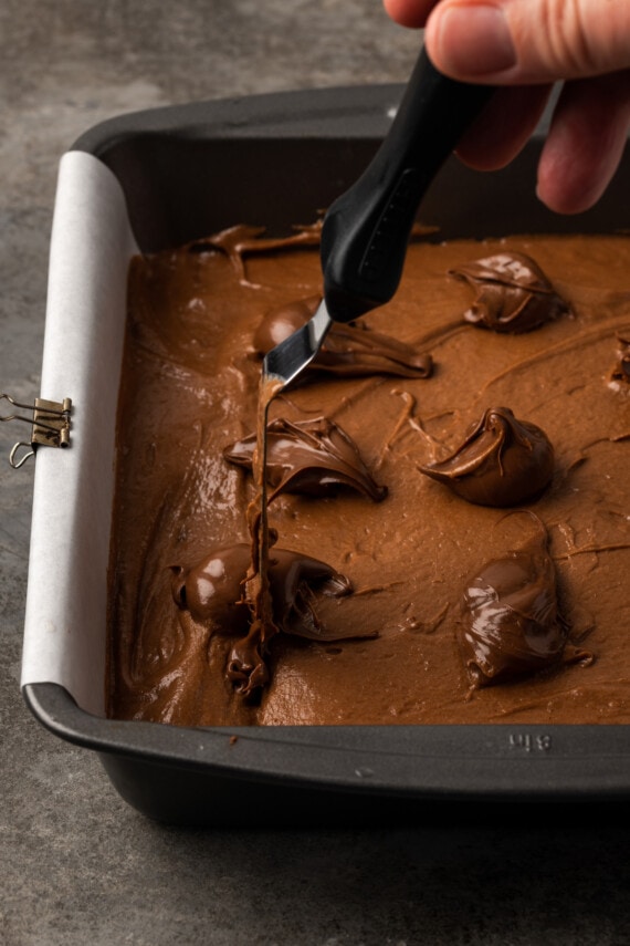Se usa una espátula desplazada para hacer girar cucharadas de Nutella en una sartén llena de masa de brownie.