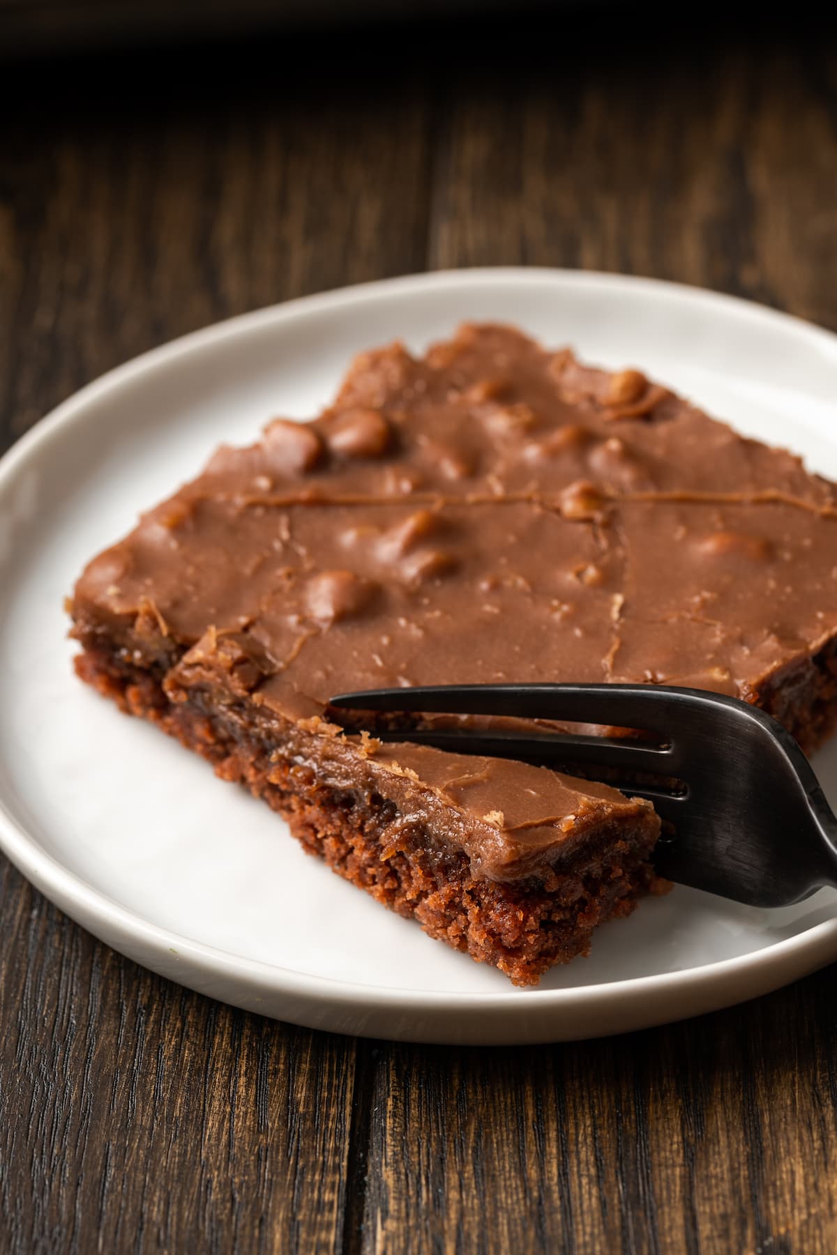 A fork digs into the corner of a slice of Texas sheet cake on a white plate.