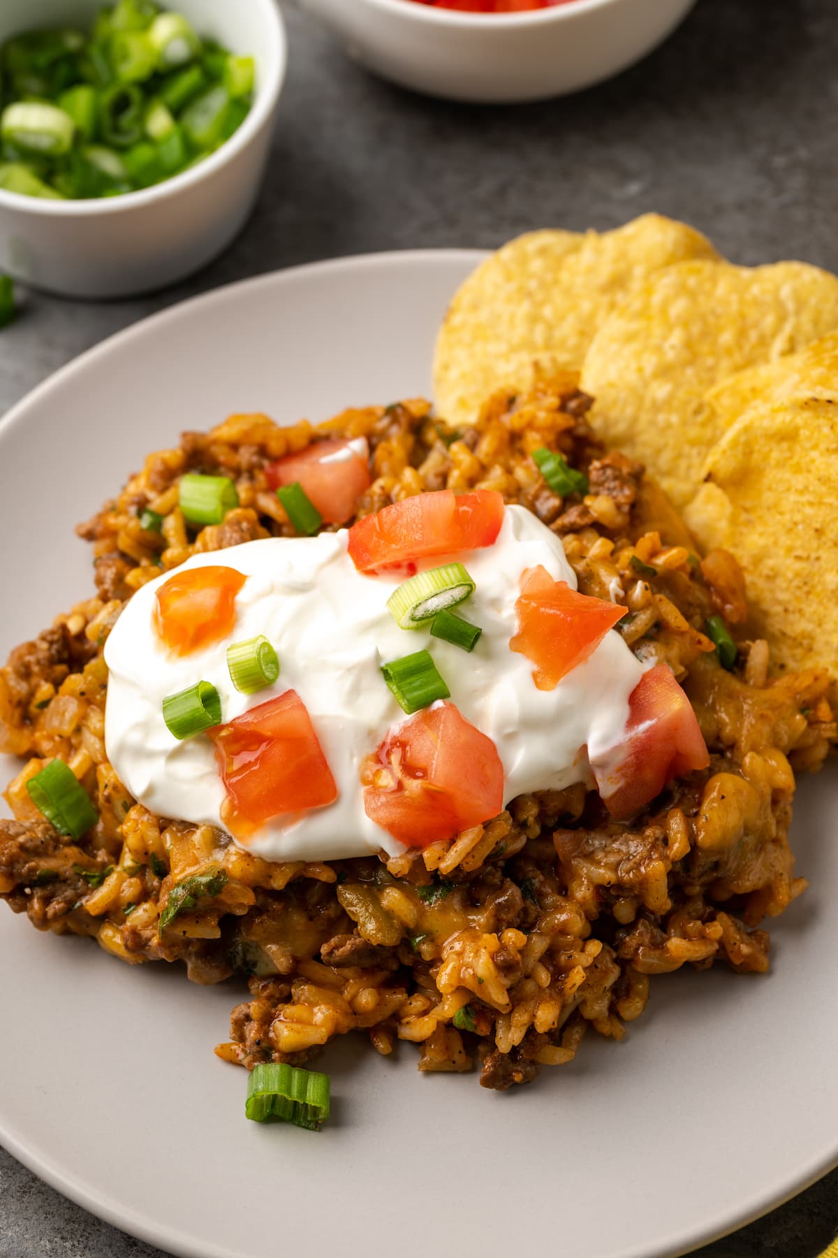 A serving of cheesy taco rice on a plate topped with sour cream, diced tomatoes, and green onions next to corn chips.