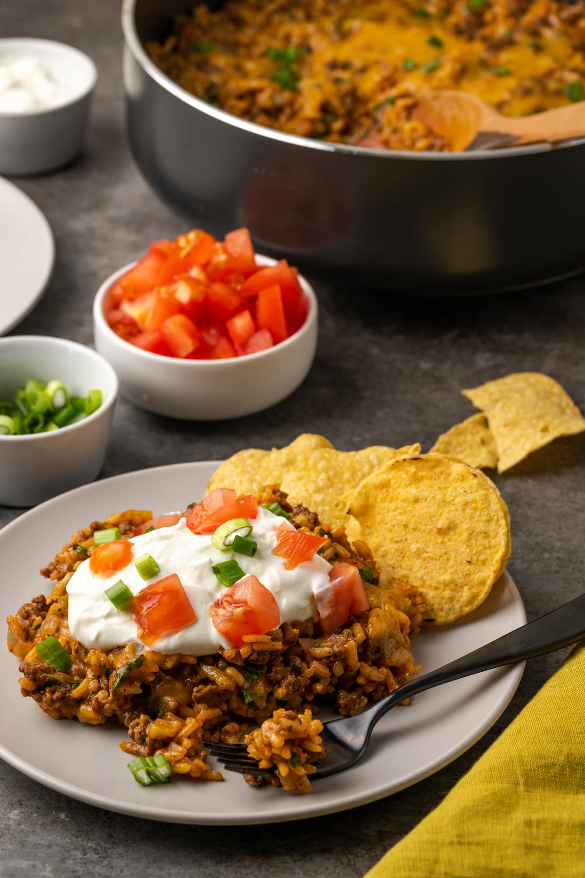 A serving of cheesy taco rice on a plate next to corn chips, topped with sour cream, diced tomatoes, and green onions with bowls of toppings and a skillet in the background.