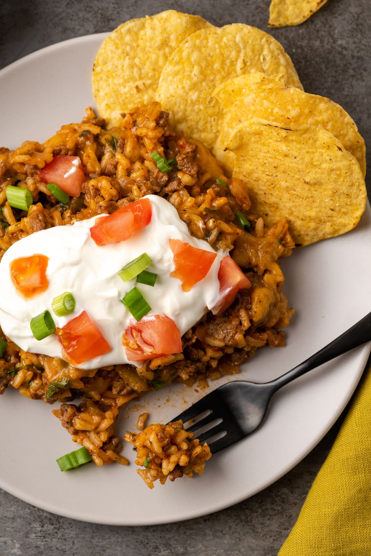 Overhead view of cheesy taco rice on a plate topped with sour cream, diced tomatoes, and green onions next to corn chips and a fork.