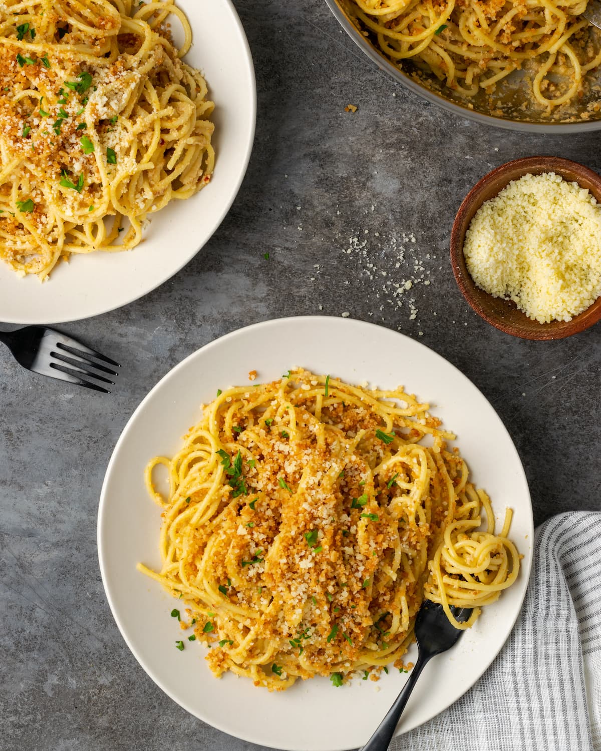 Overhead view of two platefuls of Caesar pasta topped with garlic breadcrumbs, next to a skillet and a bowl of grated parmesan cheese.