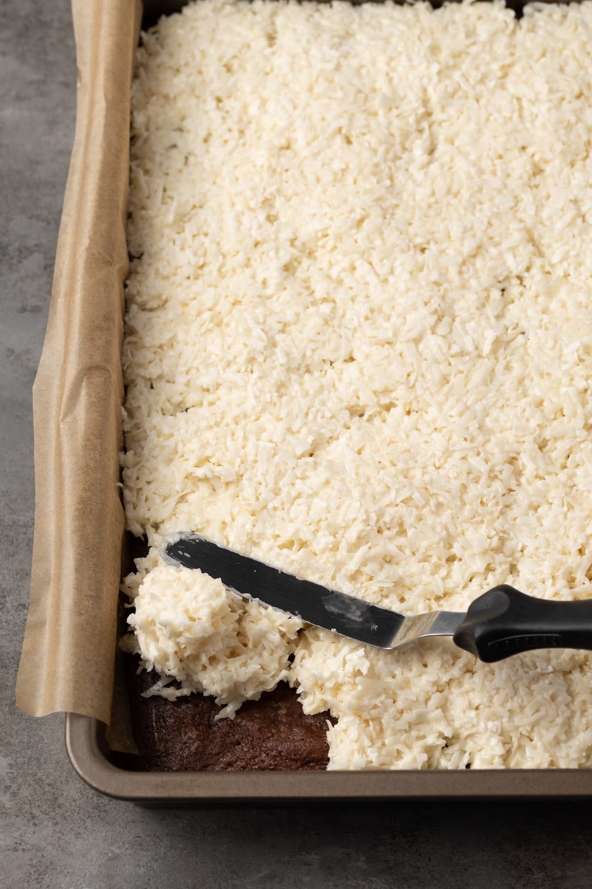 An offset spatula spreads the coconut layer over the brownies in a parchment-lined baking pan.
