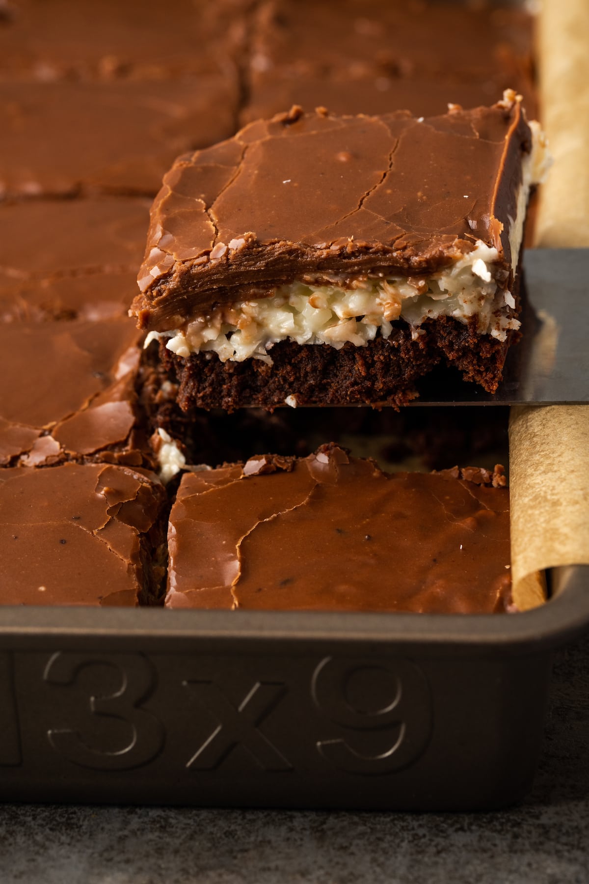 A coconut brownie is lifted from a baking pan of brownies.