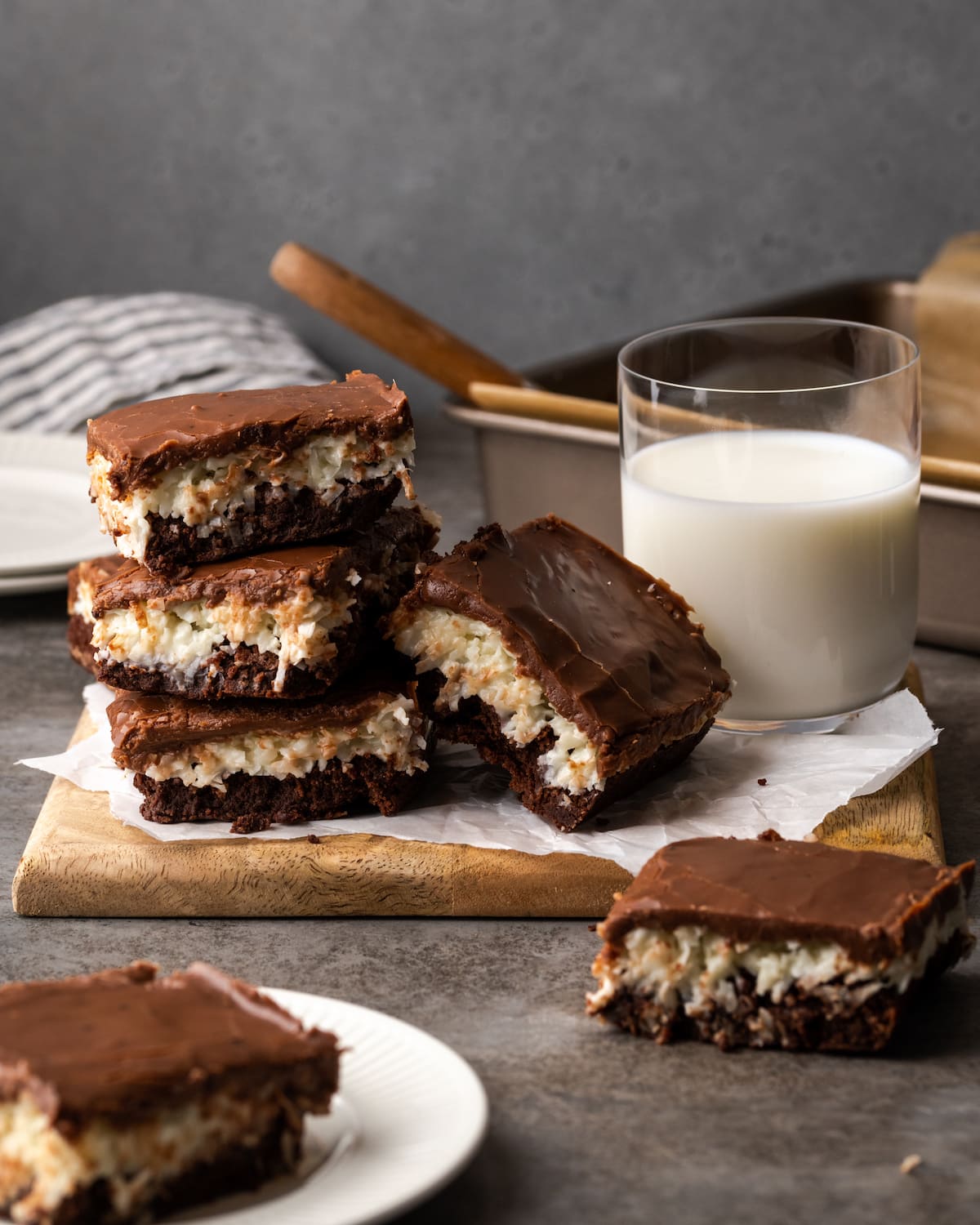 A stack of coconut brownies on a wooden platter next to a glass of milk, with a pan of brownies in the background.