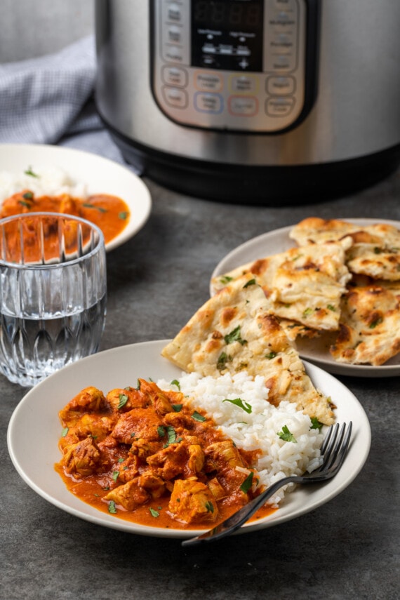 Instant pot butter chicken served on a plate next to a side of basmati rice and naan, with a plate of naan and the instant pot in the background.