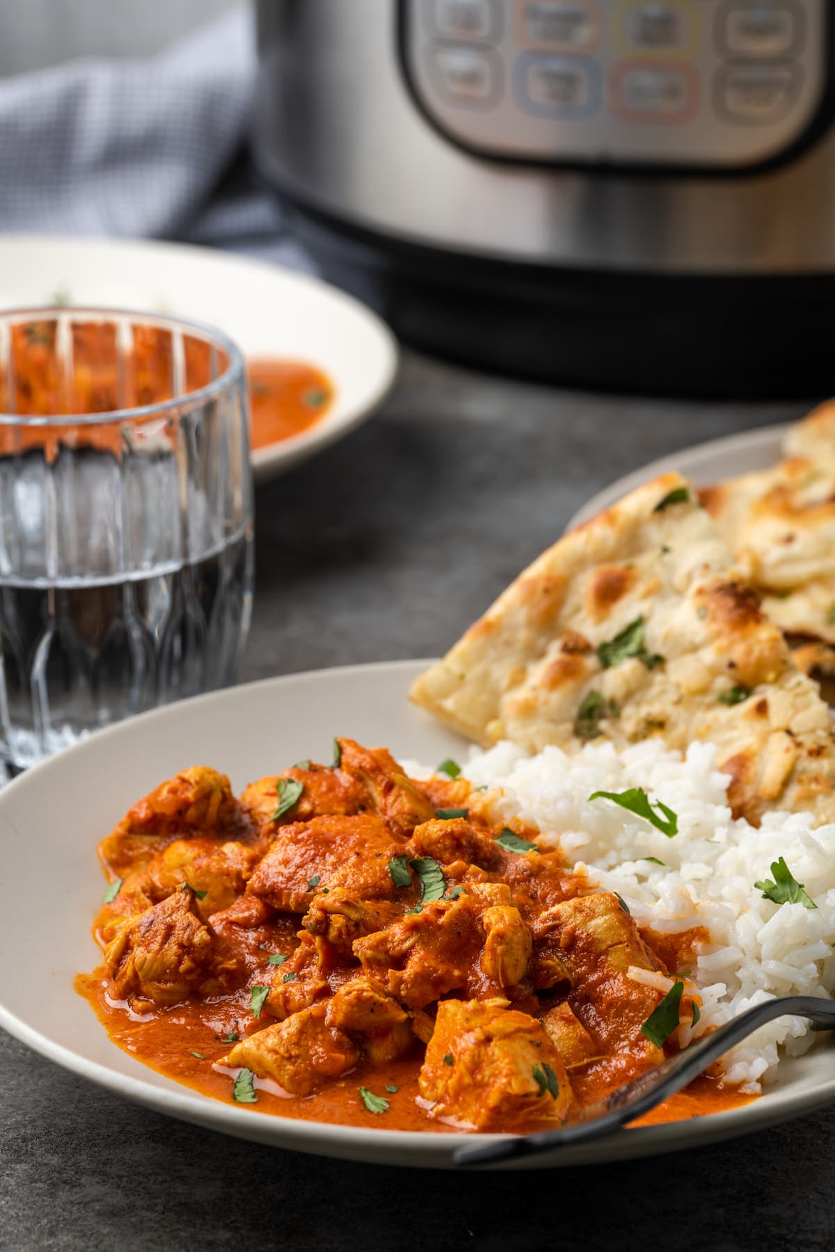 Instant pot butter chicken served on a plate next to a side of basmati rice and naan, with the instant pot in the background.