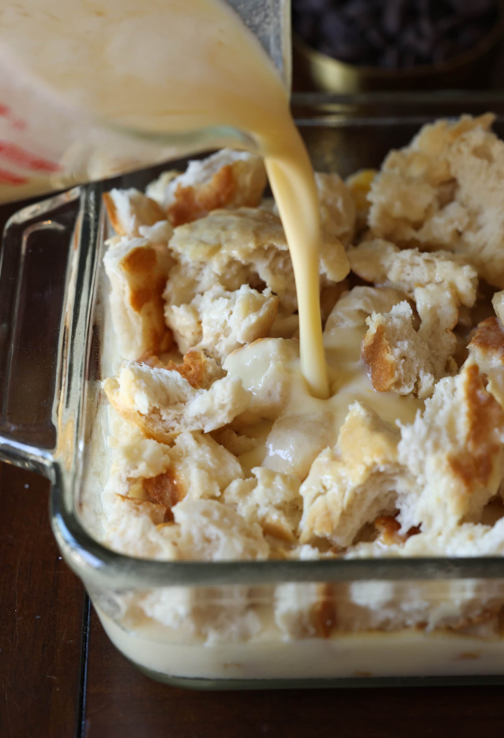 pouring a milk and egg mixture onto torn biscuit pieces in an 8x8 glass baking dish.