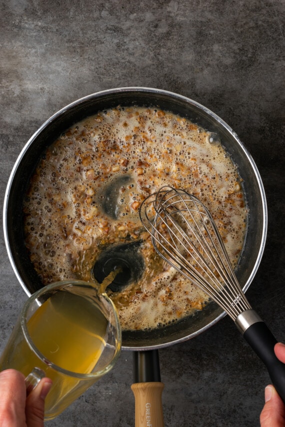 Chicken broth is whisked into a roux of flour, butter, garlic, and shallot in a skillet.
