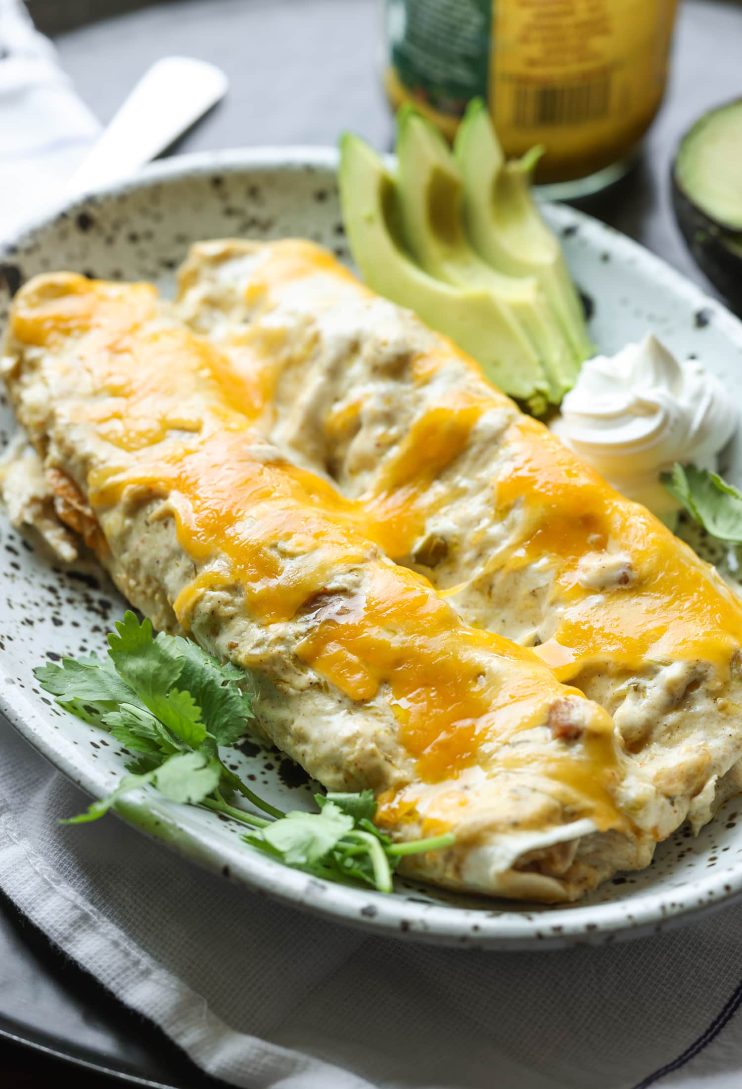 2 chicken enchiladas on a dish with cilantro, sliced avocado, and sour cream