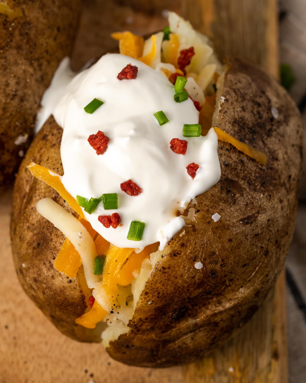 Close up of a crock pot baked potato topped with sour cream, bacon bits, and chives on a wooden board.