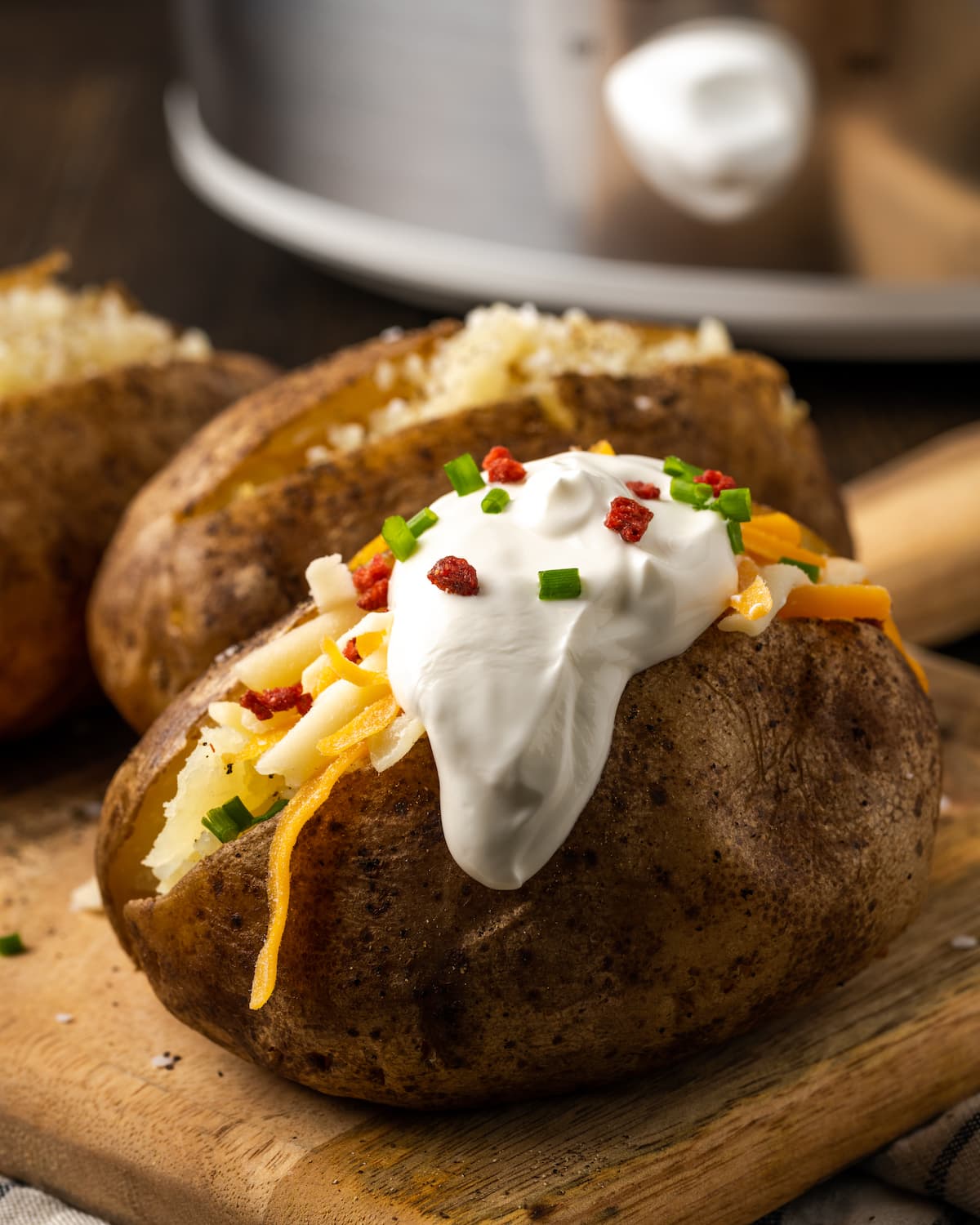 A crock pot baked potato topped with sour cream, bacon bits, and chives on a wooden board.