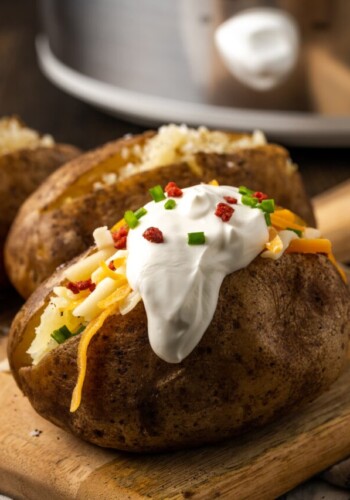 A crock pot baked potato topped with sour cream, bacon bits, and chives on a wooden board.
