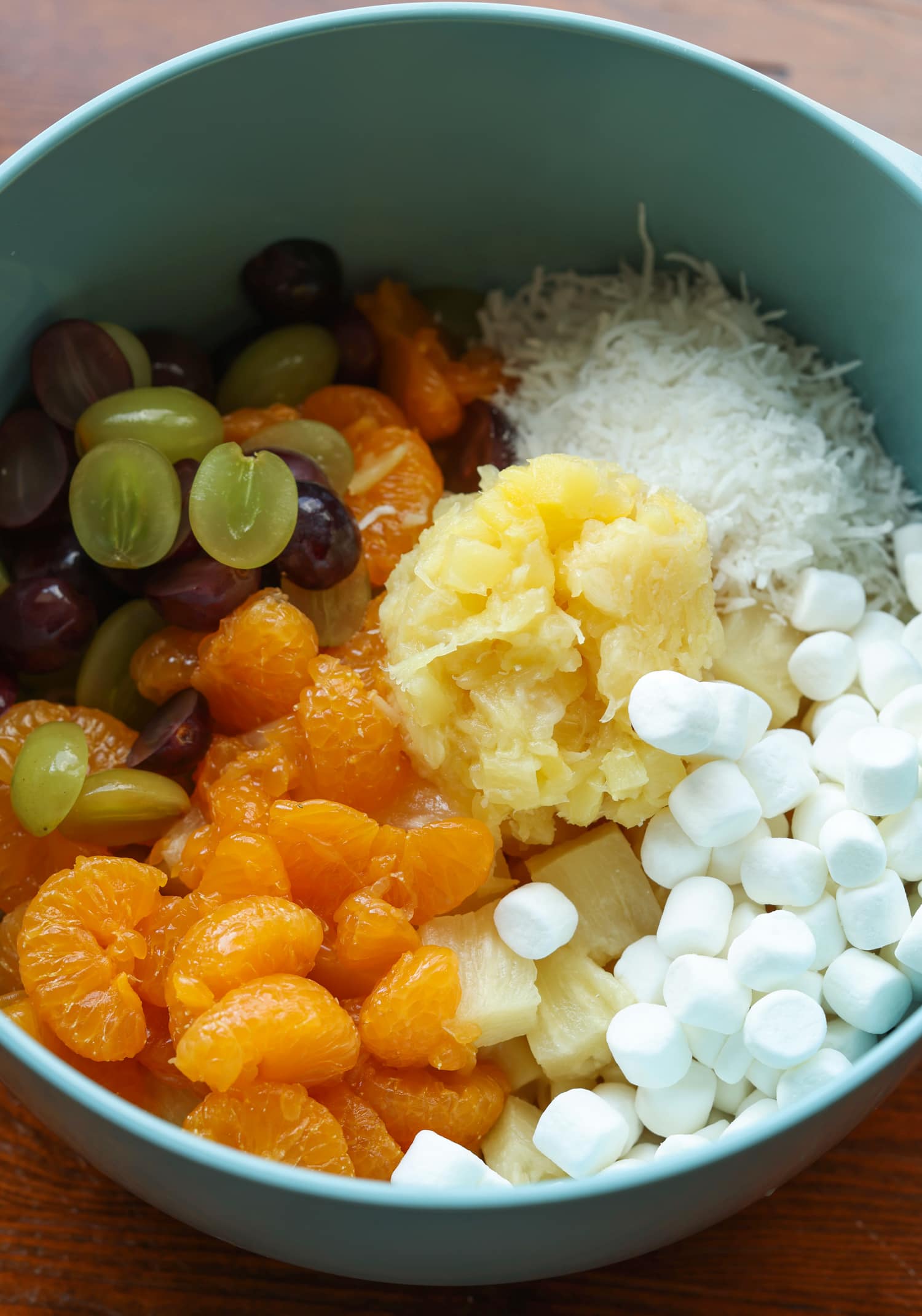 a mixing bowl with mini marshmallows, shredded coconut, crushed pineapple, mandarin oranges, and grapes.