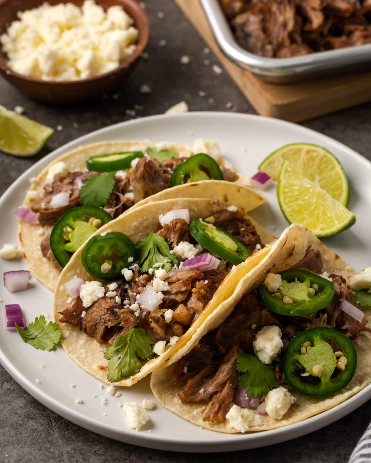 Three pork tacos topped with jalapeños and cheese on a white plate next to lime wedges.